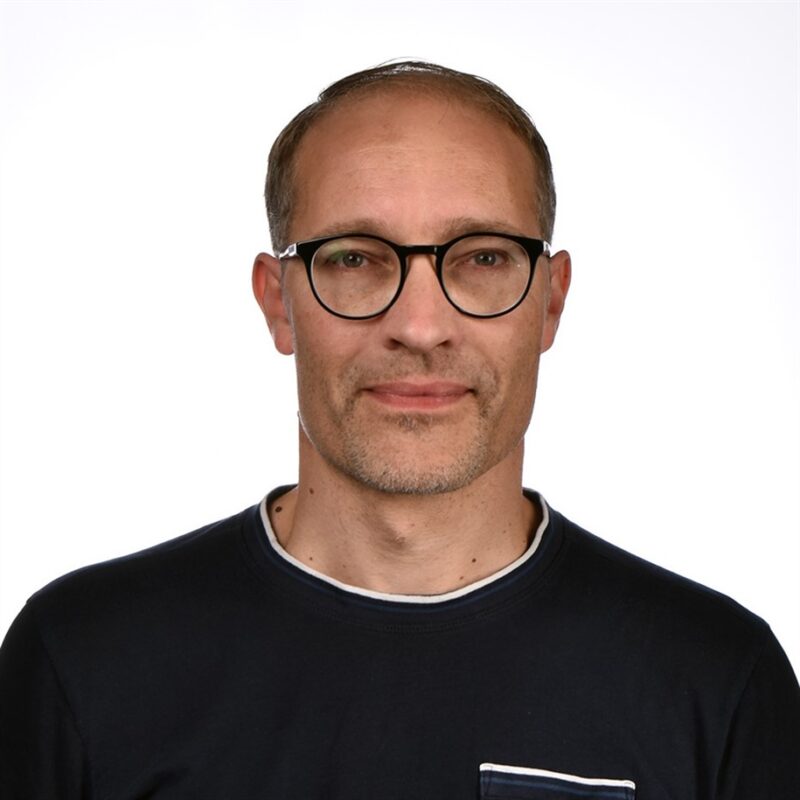 Mies, jolla on lyhyet hiukset, silmälasit ja lyhyt parta ja jolla on musta paita, tavallista valkoista taustaa vasten.