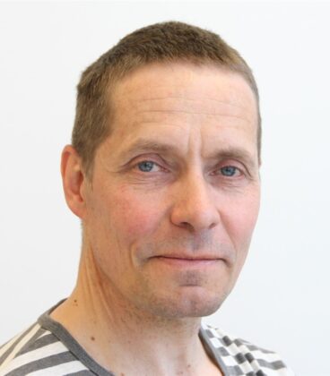 Keski-ikäinen mies, jolla on lyhyet ruskeat hiukset ja siniset silmät, katsoo kameraa neutraalilla ilmeellä yksinkertaista valkoista taustaa vasten. Hänellä on yllään valko-harmaaraidallinen paita.