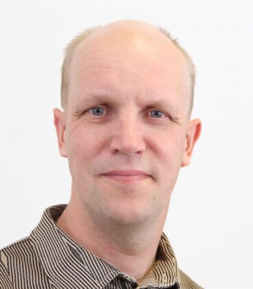 Henkilö, jolla on lyhyet, vaaleat hiukset ja neutraali ilme, katsoo suoraan kameraan. Heillä on yllään raidallinen paita ja he seisovat valkoista taustaa vasten.