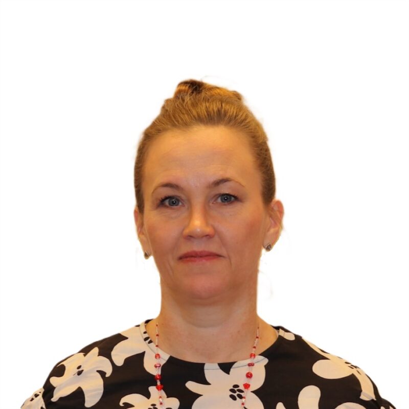 Nainen hiukset ylhäällä, yllään mustavalkoinen kukkapaita ja punaiset helmikaulakoru, seisoo valkoista taustaa vasten. Hänellä on neutraali ilme.