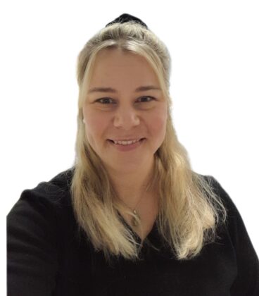 Pitkät vaaleat hiukset omaava henkilö hymyilee, yllään musta toppi ja hopeinen kaulakoru. he seisovat tavallista valkoista taustaa vasten.