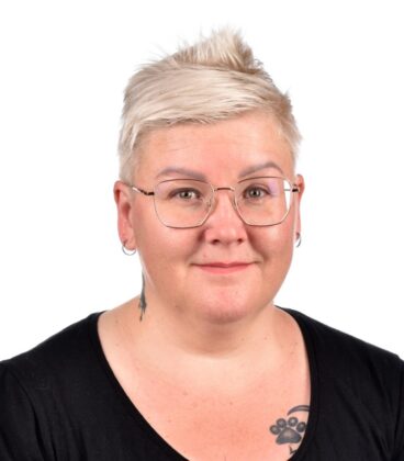 Henkilö, jolla on lyhyet, vaaleat hiukset ja silmälasit, jolla on musta toppi, poseeraa hymyillen. Heillä on tatuointi rinnassa ja pienet korvakorut. Tausta on tavallinen valkoinen.
