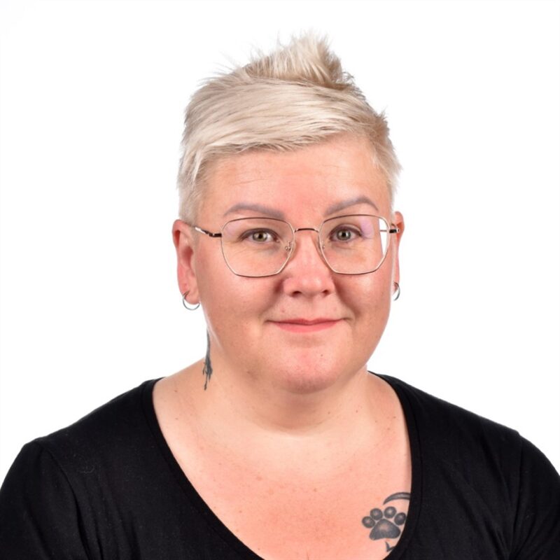 Henkilö, jolla on lyhyet, vaaleat hiukset ja silmälasit, jolla on musta toppi, poseeraa hymyillen. Heillä on tatuointi rinnassa ja pienet korvakorut. Tausta on tavallinen valkoinen.