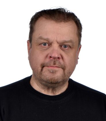Mies, jolla on lyhyet hiukset ja parta, pukeutunut mustaan paitaan, katsoo suoraan kameraan valkoista taustaa vasten.