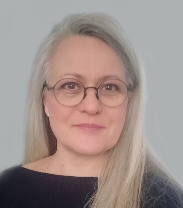 Pitkät harmaat hiukset ja pyöreät silmälasit, tumma toppi yllään, hymyilee lempeästi kameralle harmaata taustaa vasten.
