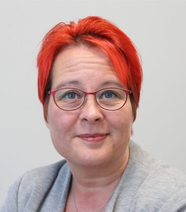 Lyhythiuksinen, kirkkaanpunainen henkilö, jolla on silmälasit ja vaaleanharmaa bleiseri, hymyilee hieman katsoessaan kameraa. Tausta on yksivärinen ja vaalea.