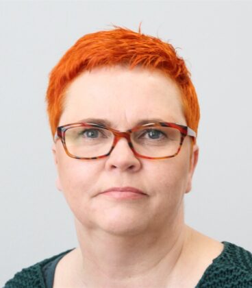 Lyhyt, kirkkaan oranssi tukka ja silmälasit omaava henkilö katsoo kameraan neutraalilla ilmeellä ja hänellä on yllään tummanvihreä paita yksiväristä vaaleaa taustaa vasten.