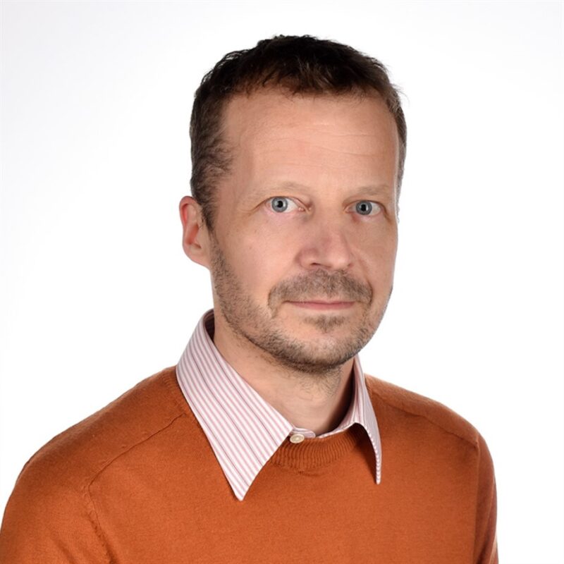Mies, jolla on lyhyet hiukset ja parta yllään oranssi pusero raidallisen paidan päällä ja katsoo kameraa valkoista taustaa vasten.