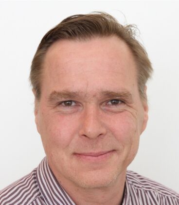 Lyhythiuksinen mies hymyilee hieman kameralle. Hänellä on yllään raidallinen paita ja hän on asetettu valkoista taustaa vasten.