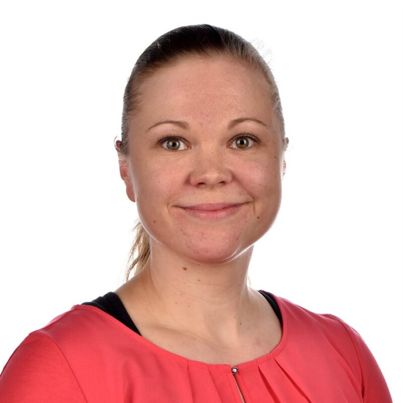 Vaaleanruskeat hiukset taakse vedetty henkilö, jolla on korallivaaleanpunainen toppi, hymyilee kameralle tavallista valkoista taustaa vasten.