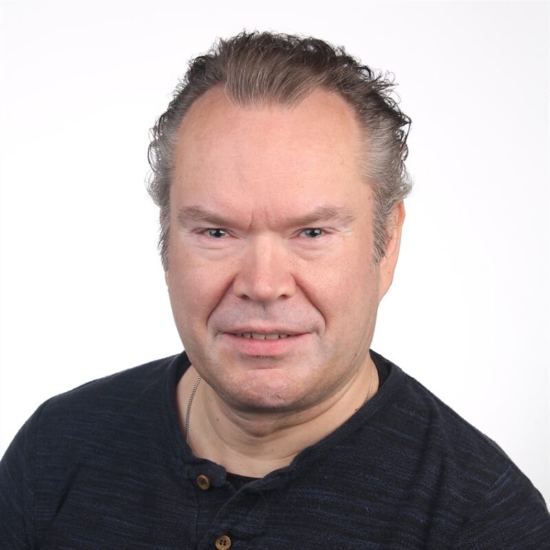 Mies, jolla on lyhyet, aaltoilevat hiukset ja yllään tumma paita, hymyilee lempeästi kameralle valkoista taustaa vasten.