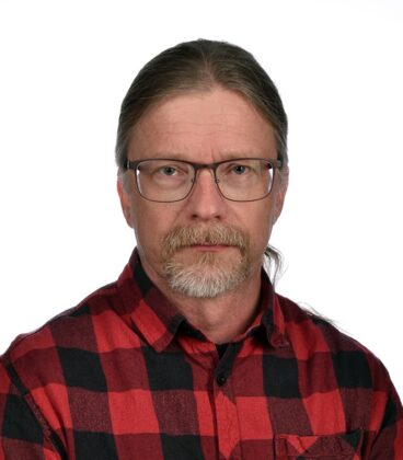 Keski-ikäinen mies, jolla on pitkät, taakse sidotut hiukset, silmälasit ja puna-musta ruutupaita, katsoo suoraan kameraan yksinkertaista valkoista taustaa vasten.