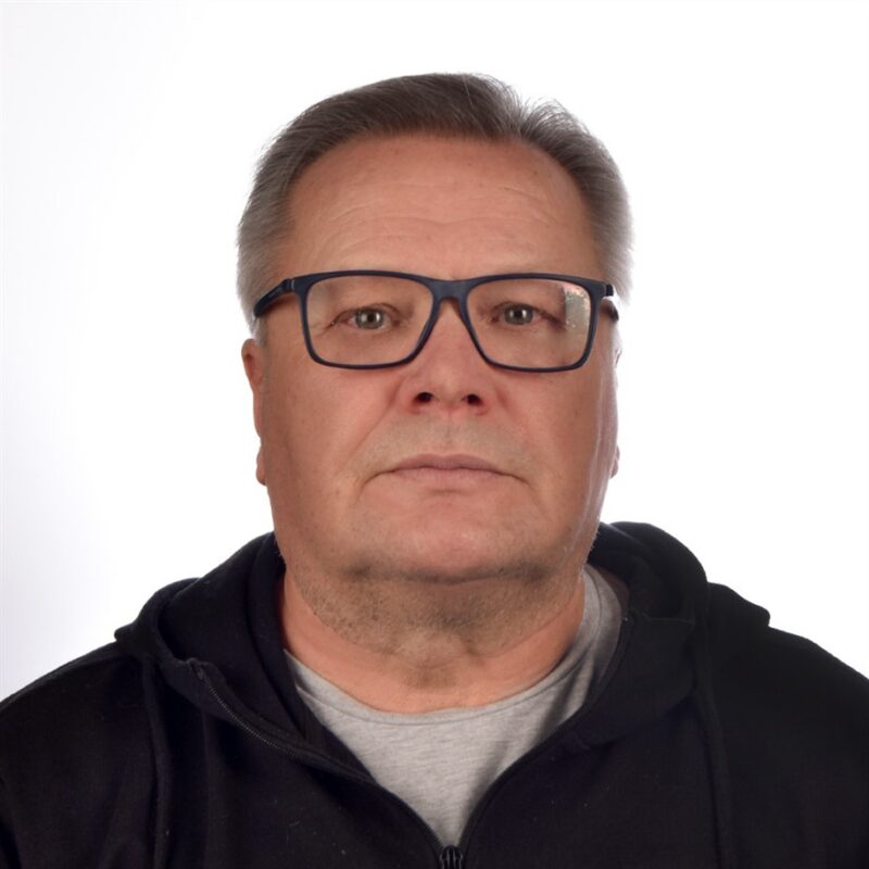 Mies, jolla on lyhyet hiukset ja silmälasit, yllään musta huppari harmaan paidan päällä ja katsoo suoraan kameraan tavallista valkoista taustaa vasten.