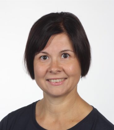 Hymyilevä lyhythiuksinen henkilö, jolla on musta paita yllään, tavallista valkoista taustaa vasten.
