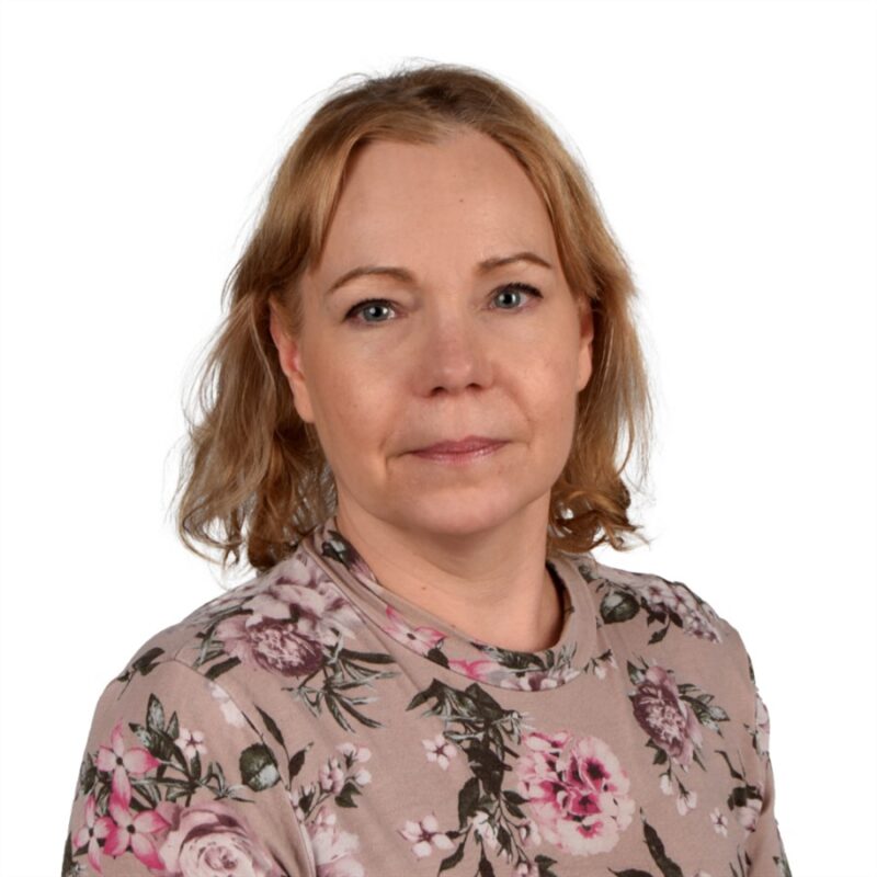 Hartioihin ulottuvilla vaaleanruskeilla hiuksilla on yllään kukkakuvioinen toppi. He katsovat suoraan kameraan neutraalilla ilmeellä. Tausta on tavallinen valkoinen.