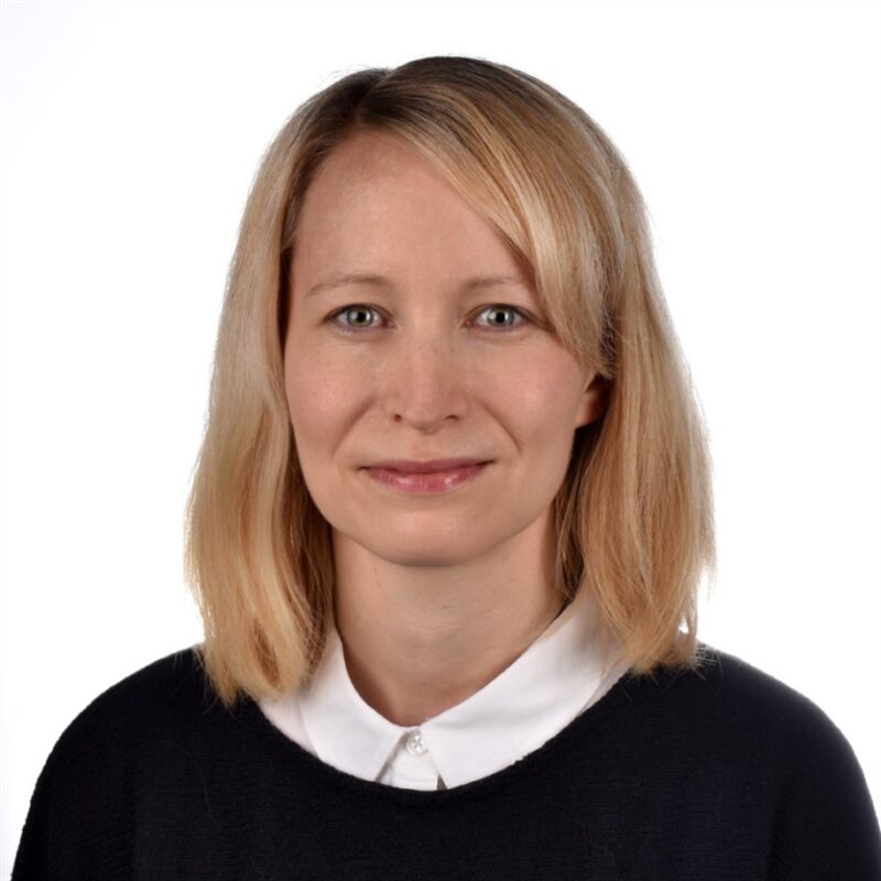 Mies, jolla on olkapäille ulottuvat vaaleat hiukset, hymyilee hieman kameralle. Heillä on yllään musta villapaita valkoisen kauluspaidan päällä. Tausta on tavallinen valkoinen.