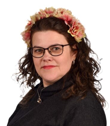 Naisella, jolla on kiharat hiukset ja silmälasit, on yllään kukkainen otsanauha ja tumma villapaita. Hän katsoo suoraan kameraan kevyesti hymyillen. Tausta on tavallinen valkoinen.