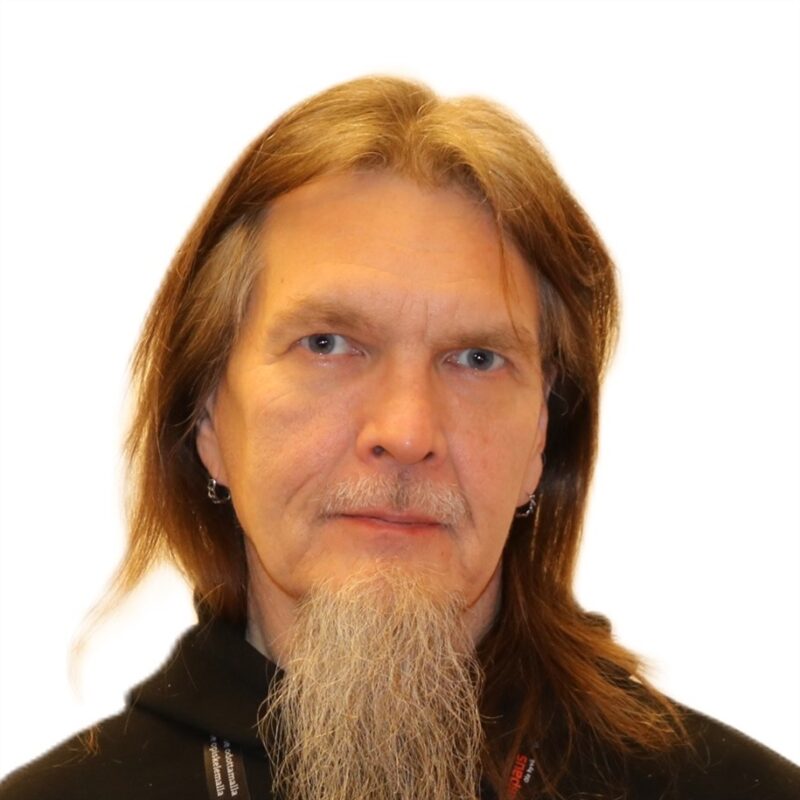 Henkilö, jolla on pitkät hiukset ja pitkä parta ja jolla on musta huppari, on katsomassa kameraa valkoista taustaa vasten. Heillä on korvakorut ja neutraali ilme.