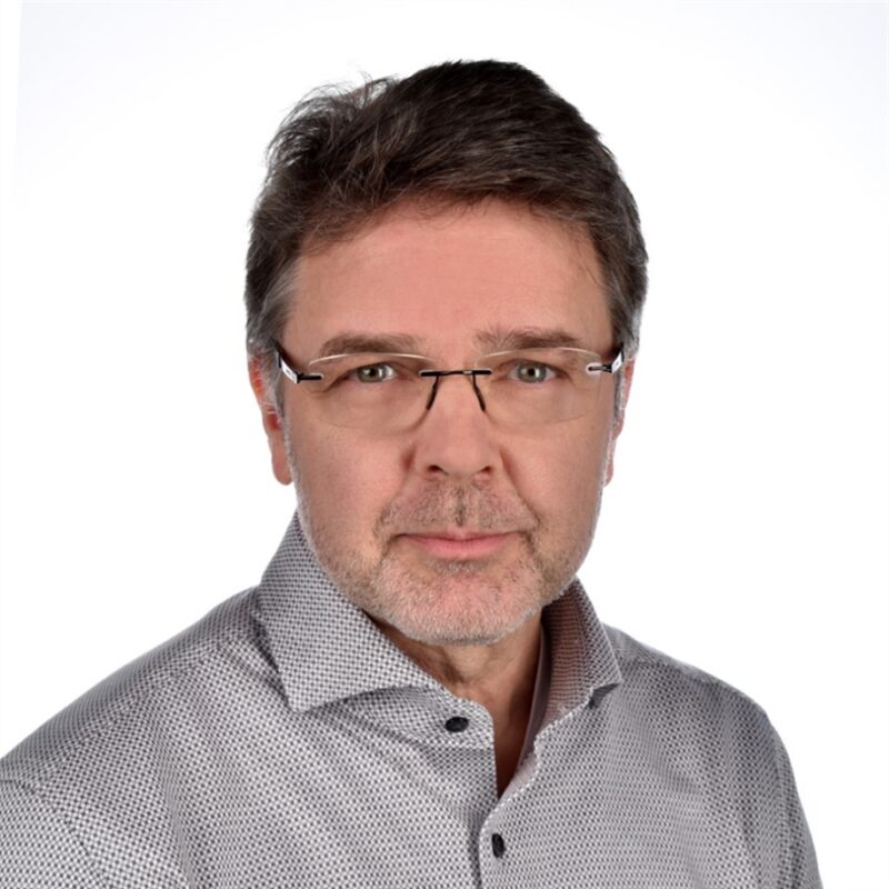 Mies, jolla on lyhyet hiukset, silmälasit ja harmaakuvioinen paita yllään, katsoo suoraan kameraan valkoista taustaa vasten.