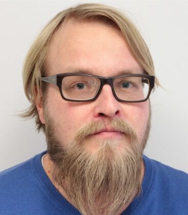 Vaaleaihoinen, vaaleatukkainen ja pitkäpartainen mies käyttää tummia suorakaiteen muotoisia laseja ja sinistä villapaitaa ja katsoo suoraan kameraan yksinkertaista vaaleaa taustaa vasten.