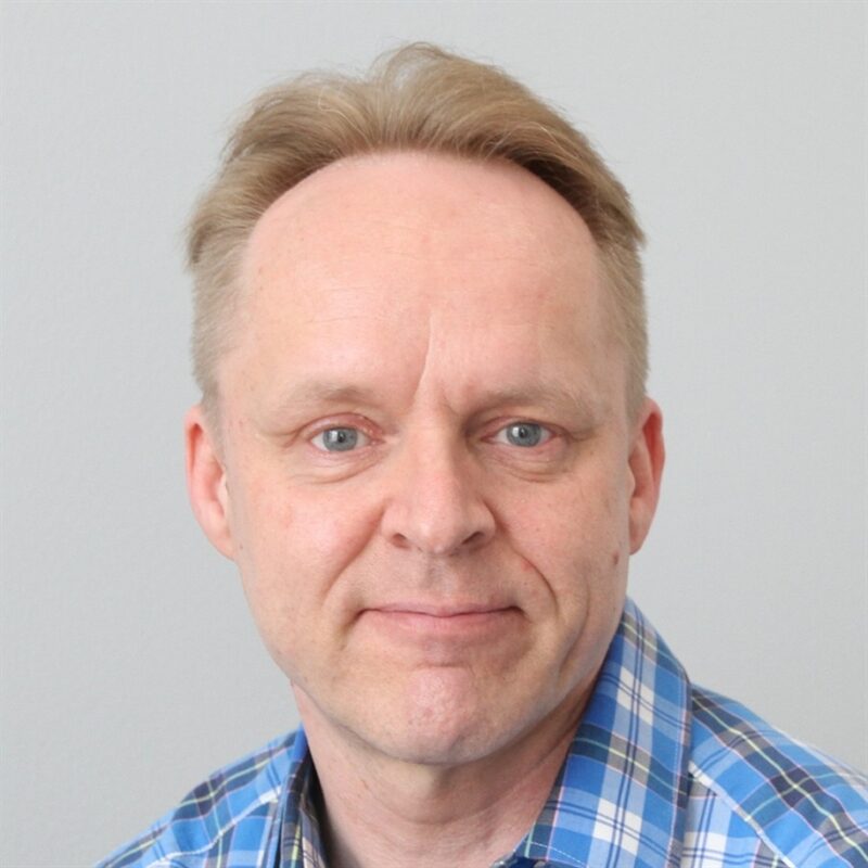 Keski-ikäinen mies, jolla on lyhyet, vaaleat hiukset ja sininen ruutupaita, hymyilee pehmeästi kameralle vaalean taustan edessä.