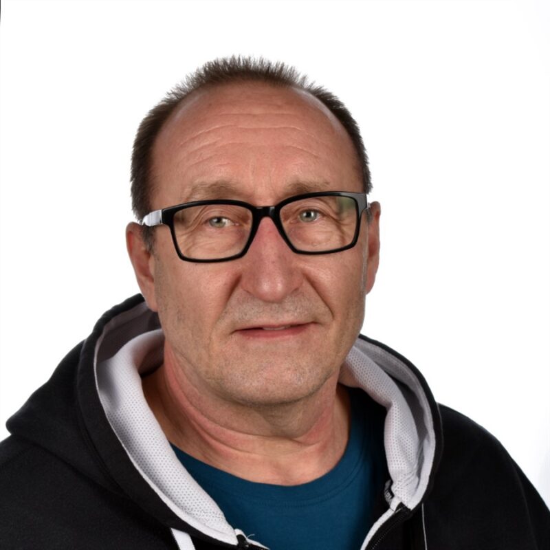 Keski-ikäisellä miehellä, jolla on lyhyet hiukset ja silmälasit, on yllään musta huppari, jossa on valkoinen sisävuori sinisen paidan päällä. Hän katsoo suoraan kameraan neutraalilla ilmeellä. Tausta on tavallinen valkoinen.