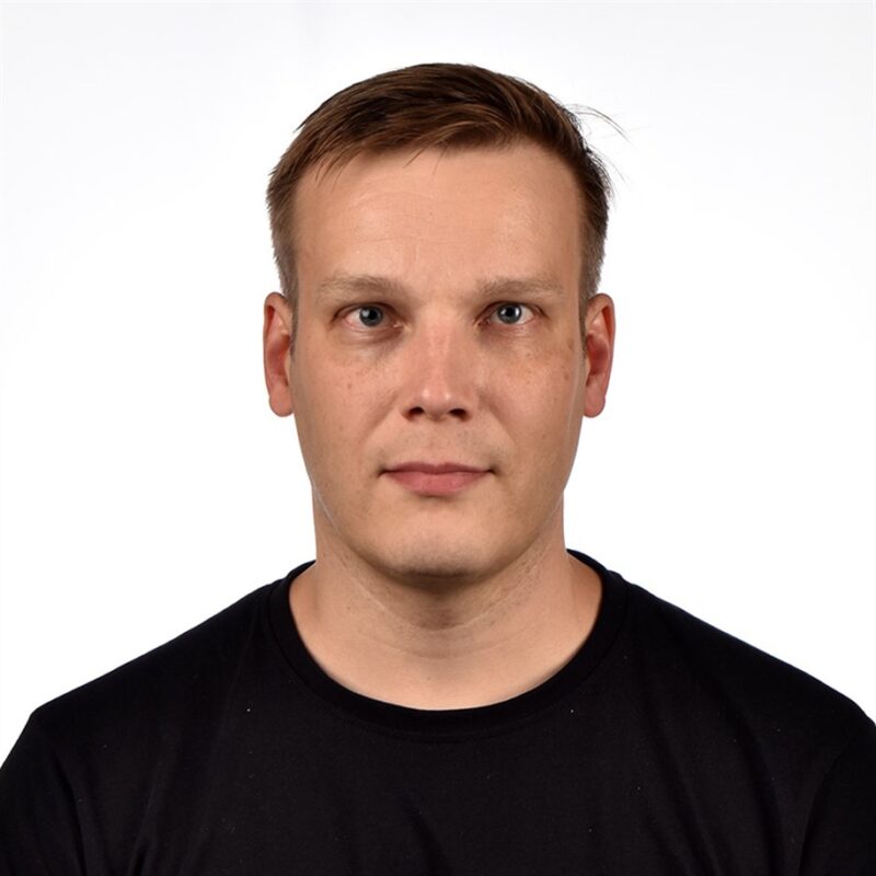 Henkilöllä, jolla on lyhyet ruskeat hiukset ja neutraali ilme, on yllään musta paita. Ne on sijoitettu valkoista taustaa vasten suoraan kameraan päin.