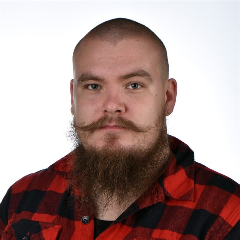 Mies, jolla on ajeltu pää, tuuhea parta ja kiharat viikset, on pukeutunut punamustaan ruutupaitaan. Hän katsoo suoraan kameraan yksinkertaista valkoista taustaa vasten.