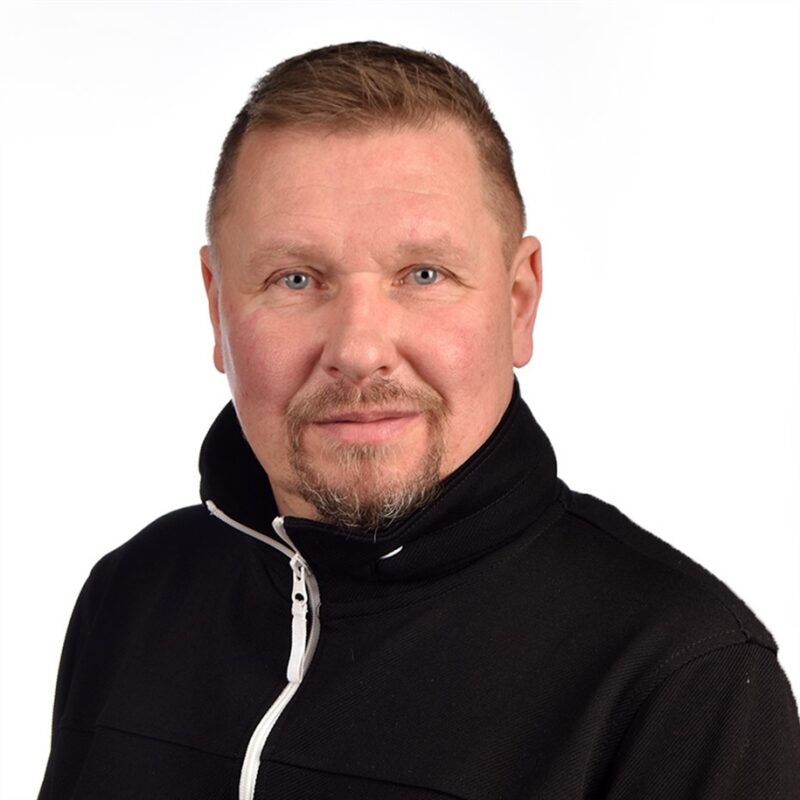 Mies, jolla on yllään musta takki, jossa on valkoinen vetoketju, hymyilee hieman kameralle tavallista valkoista taustaa vasten.