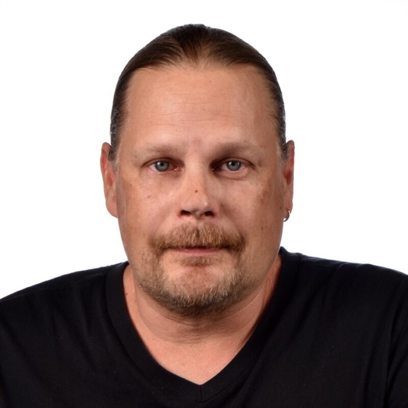 Mies, jolla on vaaleat hiukset sidottu taakse, viikset ja parta, yllään musta V-kaula-aukkoinen paita, joka katsoo suoraan kameraan neutraalilla ilmeellä valkoista taustaa vasten.