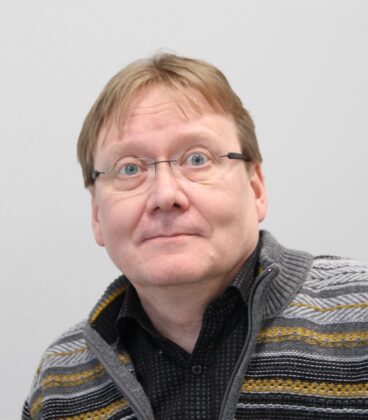 Keski-ikäinen mies, jolla on lyhyet vaaleanruskeat hiukset, silmälasit, musta paita ja harmaa kelta-valkoraitainen neulepusero, katsoo hieman ylöspäin yllättynyt ilme yksiväristä vaaleaa taustaa vasten.