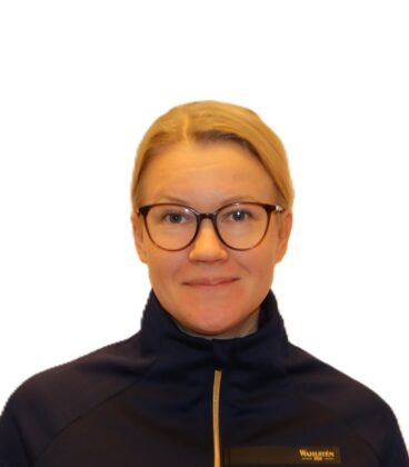 Henkilö, jolla on lyhyet vaaleat hiukset ja silmälasit, hymyilee. Heillä on yllään laivastonsininen takki, jossa on pieni logo. Tausta on valkoinen.