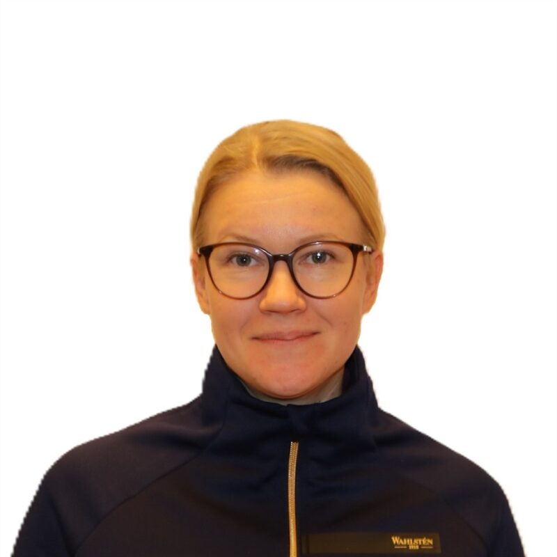 Henkilö, jolla on lyhyet vaaleat hiukset ja silmälasit, hymyilee. Heillä on yllään laivastonsininen takki, jossa on pieni logo. Tausta on valkoinen.