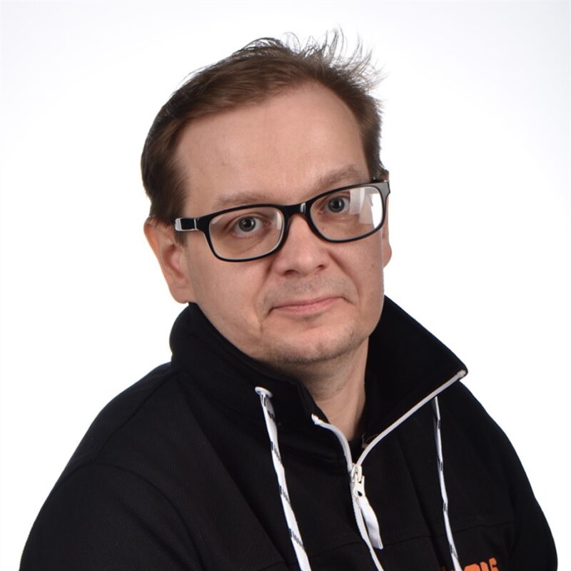 Mies, jolla on silmälasit ja musta takki valkoisella taustalla, katsoo kameraan neutraalilla ilmeellä.