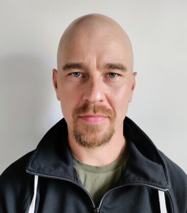 Kalju mies, jolla on vuohenpaita ja vakava ilme, yllään musta huppari vihreän paidan päällä, seisoo valkoista taustaa vasten.