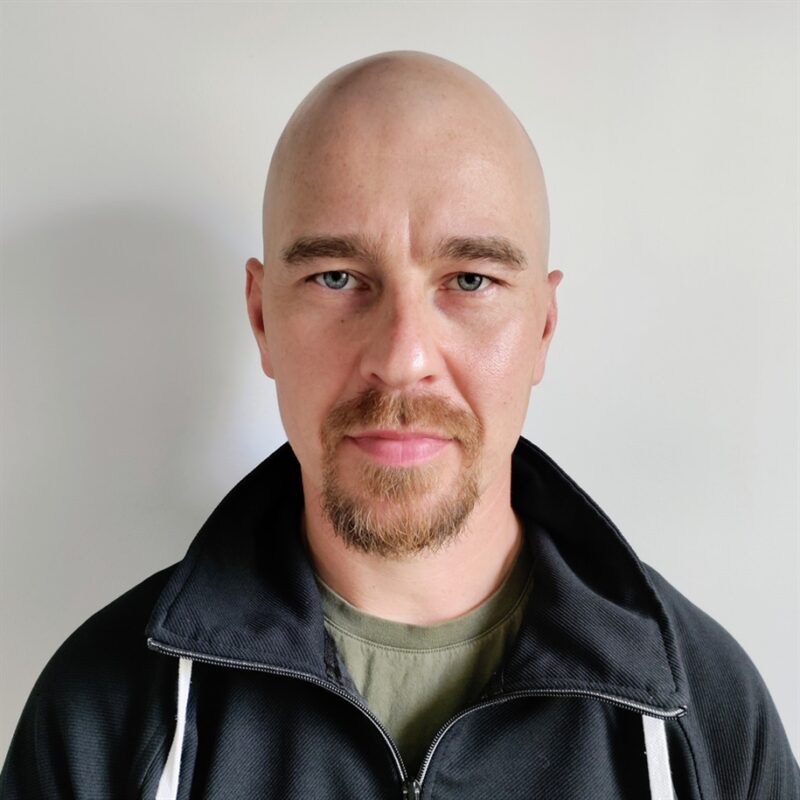 Kalju mies, jolla on vuohenpaita ja vakava ilme, yllään musta huppari vihreän paidan päällä, seisoo valkoista taustaa vasten.