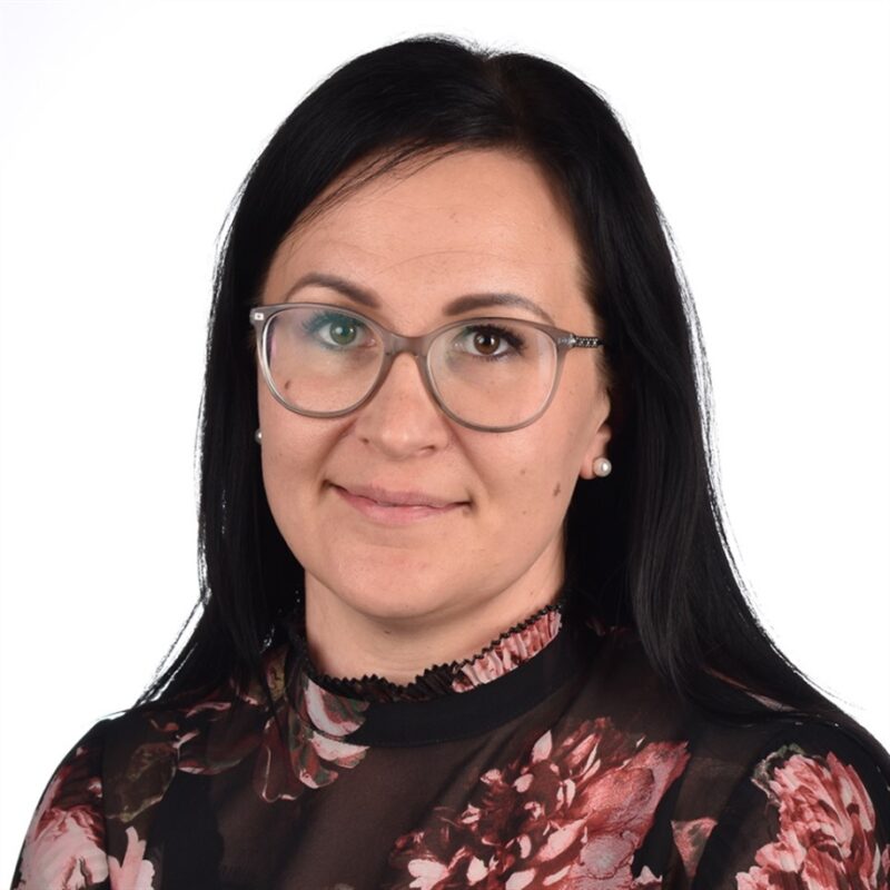 Pitkät tummat hiukset ja silmälasit pukeutunut kukkakuvioinen pusero hymyilee pehmeästi valkoista taustaa vasten.