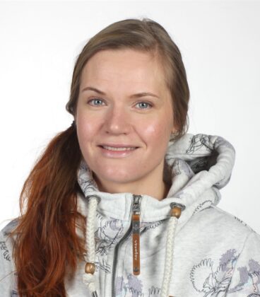 Nainen, jolla on pitkät hiukset, yllään vaalea luontokuvioinen huppari, hymyilee kameralle tavallista valkoista taustaa vasten.