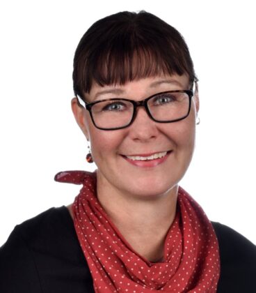 Henkilö, jolla on lyhyet tummat hiukset, hymyilee. Heillä on silmälasit, musta toppi ja punainen huivi, jossa on valkoisia pilkkuja. Tausta on tavallinen valkoinen.