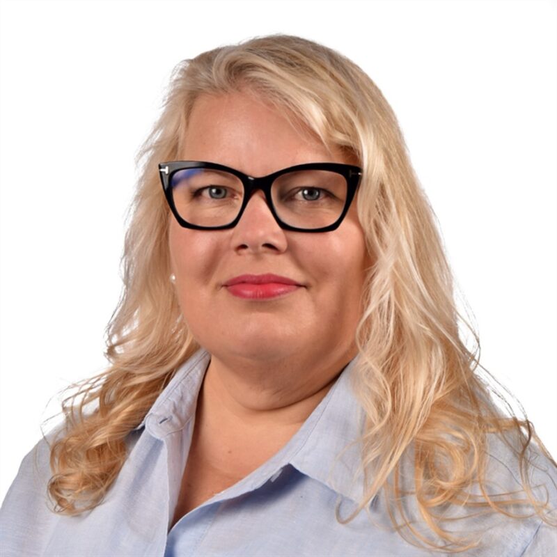 Nainen, jolla on pitkät vaaleat hiukset ja mustat lasit, yllään vaaleansininen paita, hymyilee hieman valkoista taustaa vasten.
