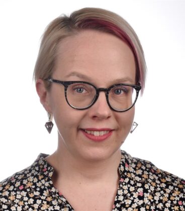 Lyhyet vaaleat hiukset ja vaaleanpunainen juova, mustat pyöreät silmälasit, geometriset korvakorut ja musta kukkakuvioinen toppi päässään hymyilevä henkilö hymyilee kameralle yksinkertaista valkoista taustaa vasten.
