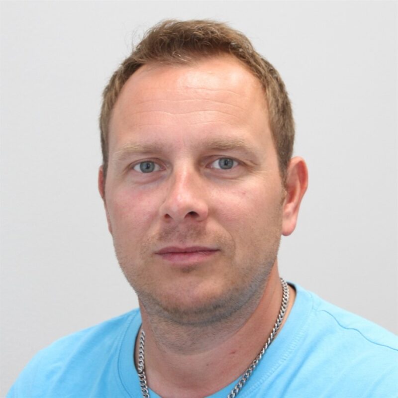 Lyhythiuksinen henkilö, jolla on vaaleansininen paita ja hopeinen kaulakoru, katsoo suoraan kameraan valkoista taustaa vasten.