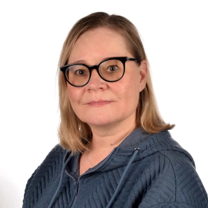 Naisella, jolla on olkapäille ulottuvat vaaleat hiukset ja mustat lasit, on yllään sininen tikattu takki. Hän katsoo suoraan kameraan tavallista valkoista taustaa vasten.