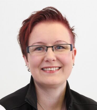 Henkilö, jolla on lyhyet, punertavat hiukset ja silmälasit, hymyilee kameralle. Heillä on yllään musta takki valkoisen paidan päällä tavallista valkoista taustaa vasten.