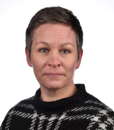 Lyhyttukkaisella henkilöllä on yllään mustavalkokuvioinen neule. He katsovat suoraan kameraan neutraalilla ilmeellä. Tausta on tavallinen valkoinen.