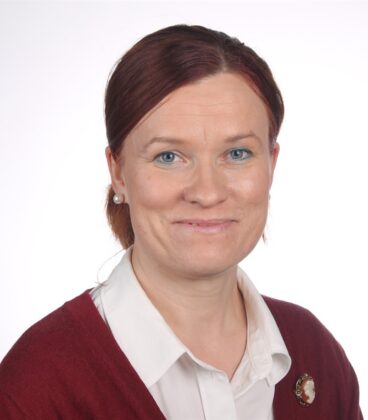 Punaiset hiukset selkään sidottulla henkilöllä on yllään valkoinen pusero ja viininpunainen villapaita. He hymyilevät ja seisovat tavallista valkoista taustaa vasten.