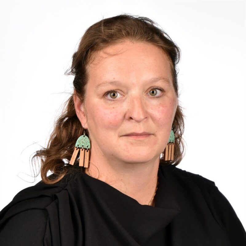 Pitkät ruskeat hiukset nainen, jolla on suuret koristeelliset korvakorut ja musta toppi, seisoo valkoista taustaa vasten. Hänellä on neutraali ilme.