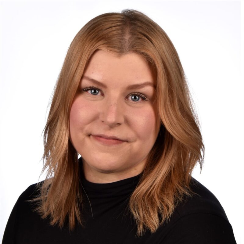 Hartioihin ulottuva, vaaleanruskeat hiukset ja musta toppi omaava henkilö hymyilee pehmeästi. Tausta on puhdas, kirkkaan valkoinen.