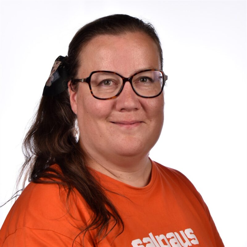 Pitkät ruskeat hiukset omaava nainen, jolla on suuret mustat silmälasit, oranssi paita ja musta rusetti, hymyilee kameralle valkoista taustaa vasten.