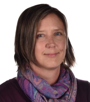 Lyhythiuksinen henkilö, jolla on violetti paita ja värikäs kuviollinen huivi, hymyilee hieman tavallisen valkoisen taustan edessä.